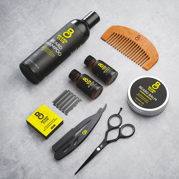Beard Club grooming kit with shampoo, oil, balm, wooden comb, scissors, and a straight razor on a grey surface.
