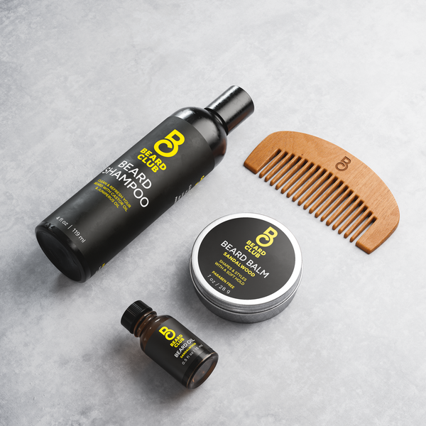 Beard Club kit with shampoo, sandalwood balm, oil, and a wood comb on a grey surface.
